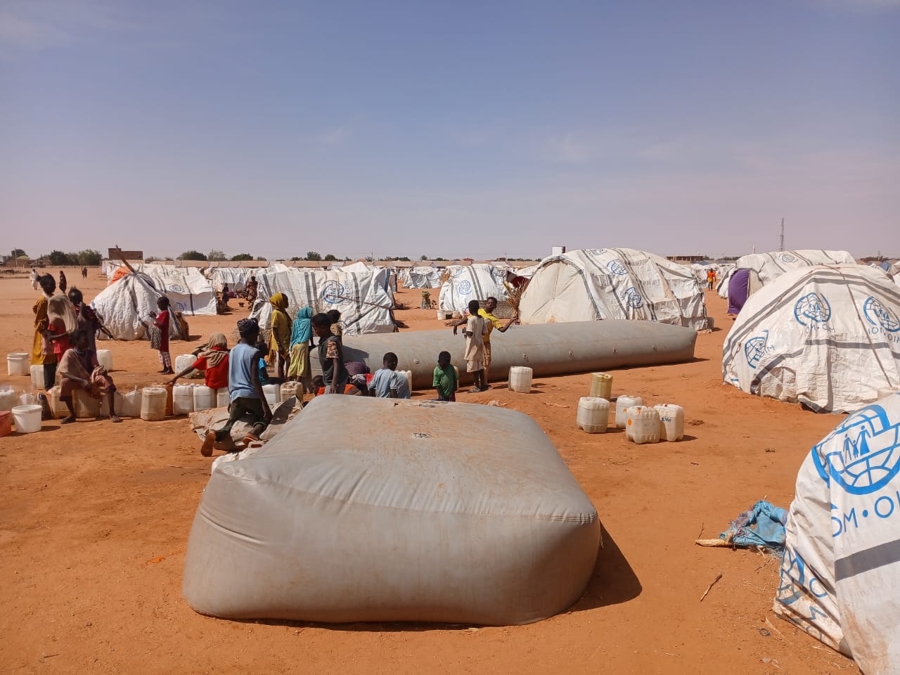 Wide shot of displaced people gathered around a water bladder near their plastic-sheet tents in Al-Mina Al Muwahad, the largest displacement camp near El Obeid city, hosting approximately 25,000 people.