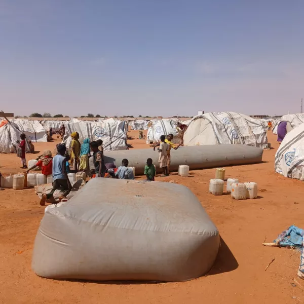 Wide shot of displaced people gathered around a water bladder near their plastic-sheet tents in Al-Mina Al Muwahad, the largest displacement camp near El Obeid city, hosting approximately 25,000 people.