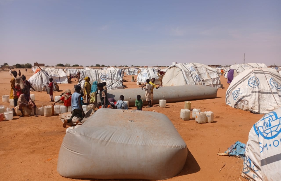 Wide shot of displaced people gathered around a water bladder near their plastic-sheet tents in Al-Mina Al Muwahad, the largest displacement camp near El Obeid city, hosting approximately 25,000 people.