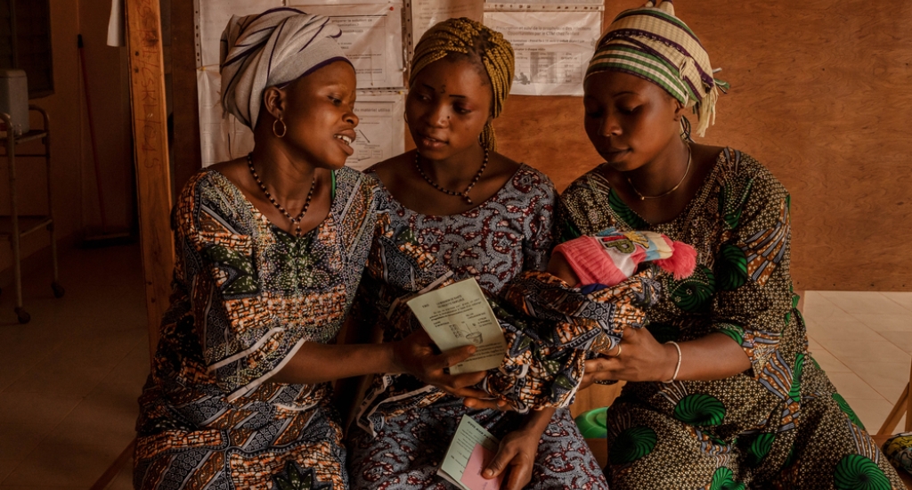 Hortense (links) geeft haar pasgeboren dochter aan Mélanie (rechts), die zes maanden zwanger is, terwijl ze wachten op een consult in het gezondheidscentrum van Klouékanmè. Hortense beviel drie dagen eerder alleen thuis van haar dochter. Couffo, Benin, januari 2025.