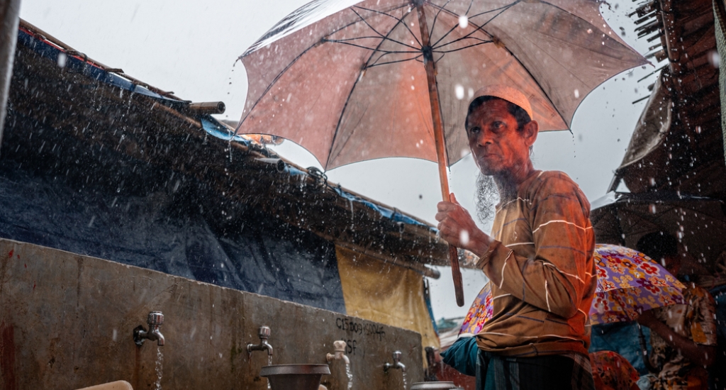 Ershadullah, een Rohingya-vluchteling die in 2017 uit Myanmar vluchtte, zoekt beschutting onder een paraplu tijdens hevige regen bij een AZG-waterpunt in het vluchtelingenkamp Kutupalong. Cox’s Bazar, Bangladesh, mei 2025.